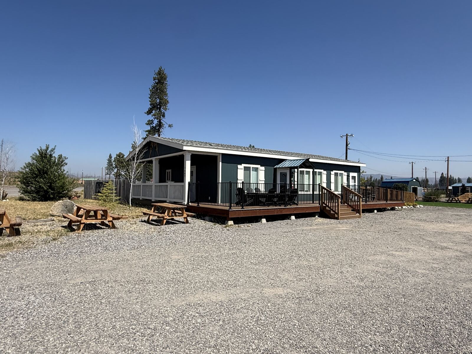 The house at Cedar Lodge with its covered porch and long railed deck, picnic tables on the gravel in front and a tall pine standing behind it.